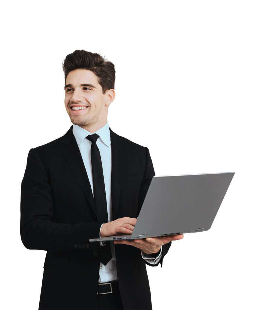 A man wearing a black suit and tie, is smiling while holding and using a laptop.